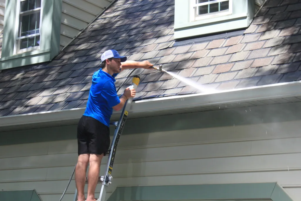 AllPro technician on a ladder cleaning the gutters and roofline of a green-sided home