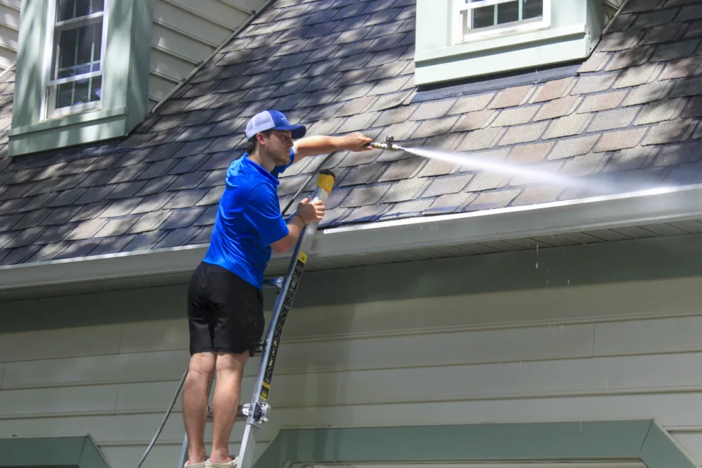 AllPro technician on a ladder cleaning the gutters and roofline of a green-sided home